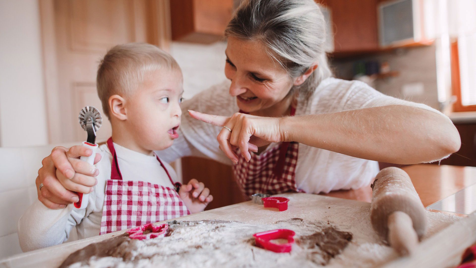 A happy handicapped down syndrome child and his mother with checked aprons indoors baking in a kitchen, having fun.